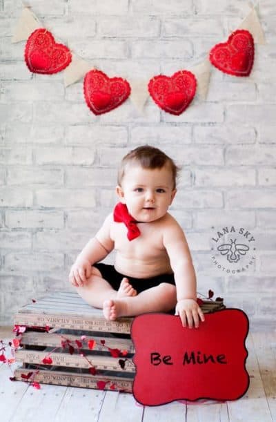 Baby surrounded by heart decor holding sign that says be mine