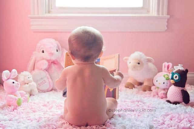 Baby sitting in bedroom reading book to stuffed animals