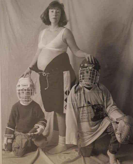 Mom poses in hockey pants and sports bra while fully clothed children in hockey gear crouch beneath her.