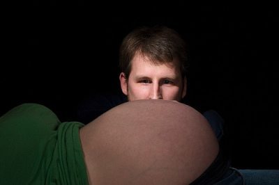 Forefront of photo shows woman laying on her back in the shadows with her bump exposed, while a man's lit up face is shown staring at the bump. 