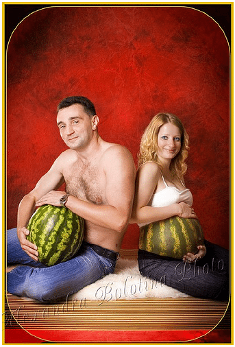 Couple sits back to back smiling at camera. He is topless cradling a watermelon, she is wearing a white tank top and has her bump painted like a watermelon.