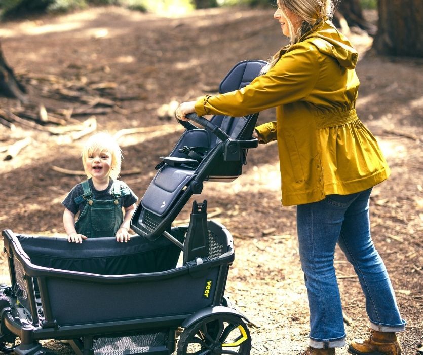 Woman attaching Veer Switchback seat to Veer Cruiser wagon while smiling toddler watches.