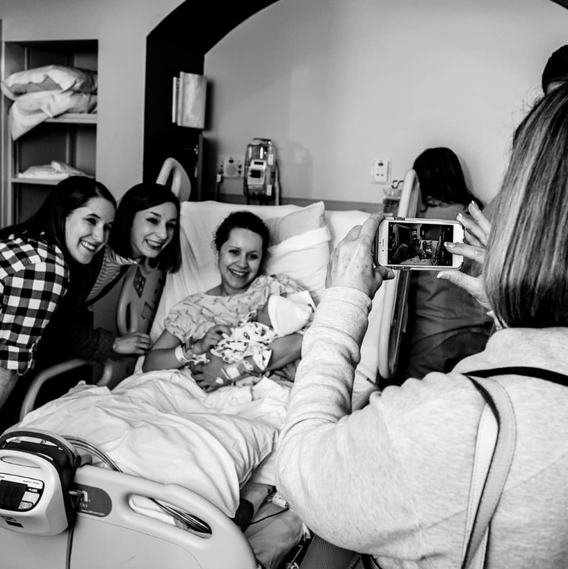 Black and white photo of new mom holding baby while two sisters stand next to her and adoring new grandma takes a photo with her phone.