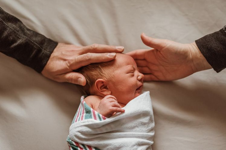 Color image of baby sleeping on a white sheet, swaddled, while mom and dad each but a hand under the head.