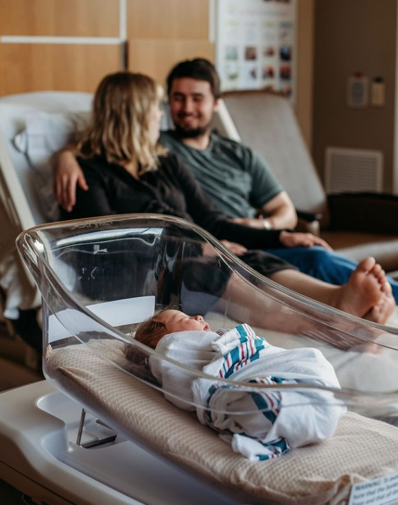 Color photo of swaddled baby in hospital bassinet while parents are in the background smiling at each other. 