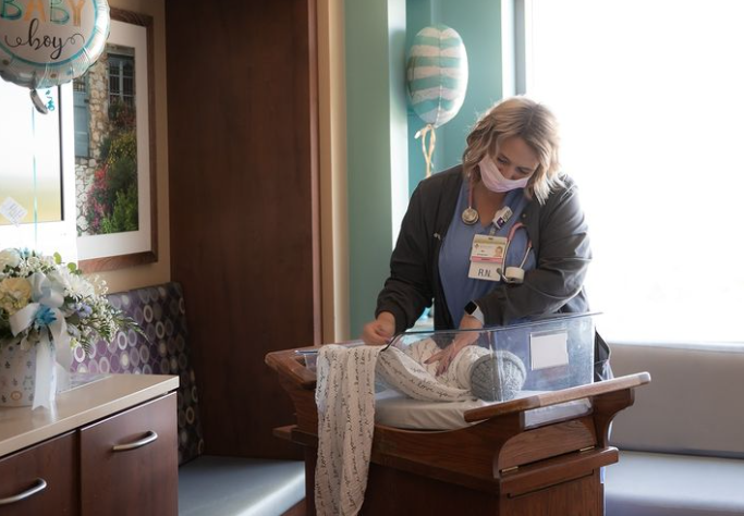 Color photo of nurse looking adoringly at swaddled baby in hospital bassinet. 