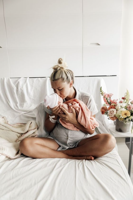 Color photo of mom kissing baby swaddled in pink next to bouquet of pink, yellow and white flowers.