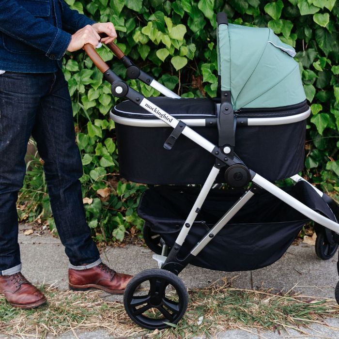 Parent pushing Mockingbird single stroller with bassinet attached on sidewalk with green ivy wall behind.