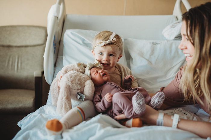 Toddler big sister holding crying newborn baby sister sitting on hospital bed