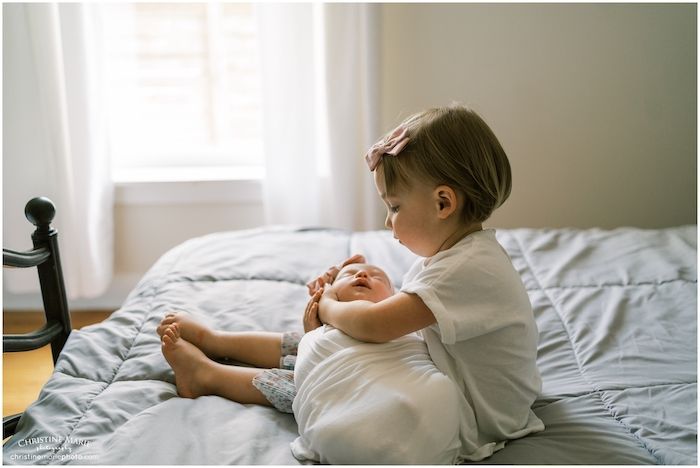 Toddler big sister holding newborn baby sibling at home on bed