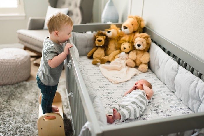 Toddler big brother standing on stool to look at new baby in crib