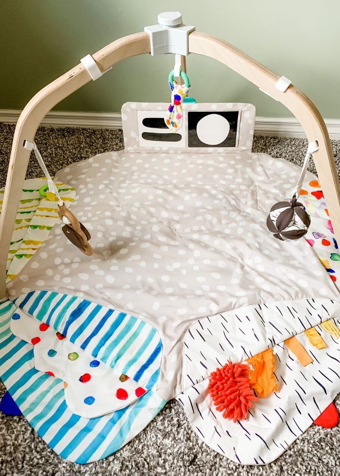 Photo of Lovevery play gym set up and unfolded on the floor. The 5 zones of play are open exposing different flaps, colors, textures, and cards. The wooden batting right, organic teether, and Montissori ball all hang from the wooden legs.