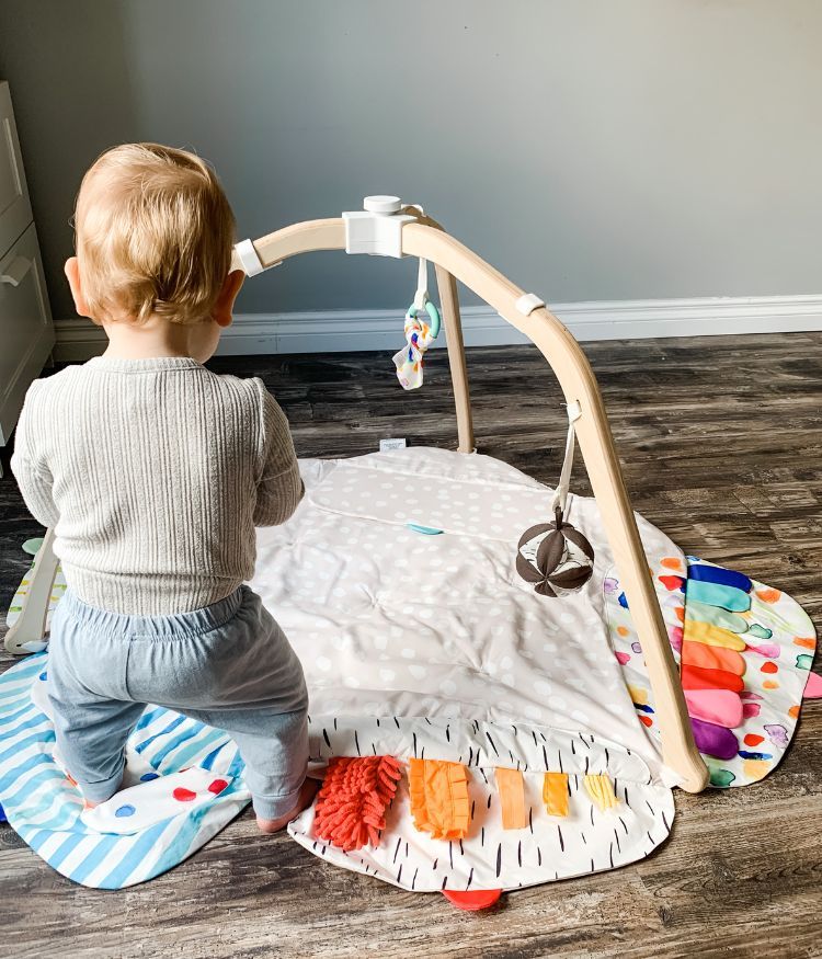 Baby standing near Lovevery play gym inspecting one of the hanging toys.